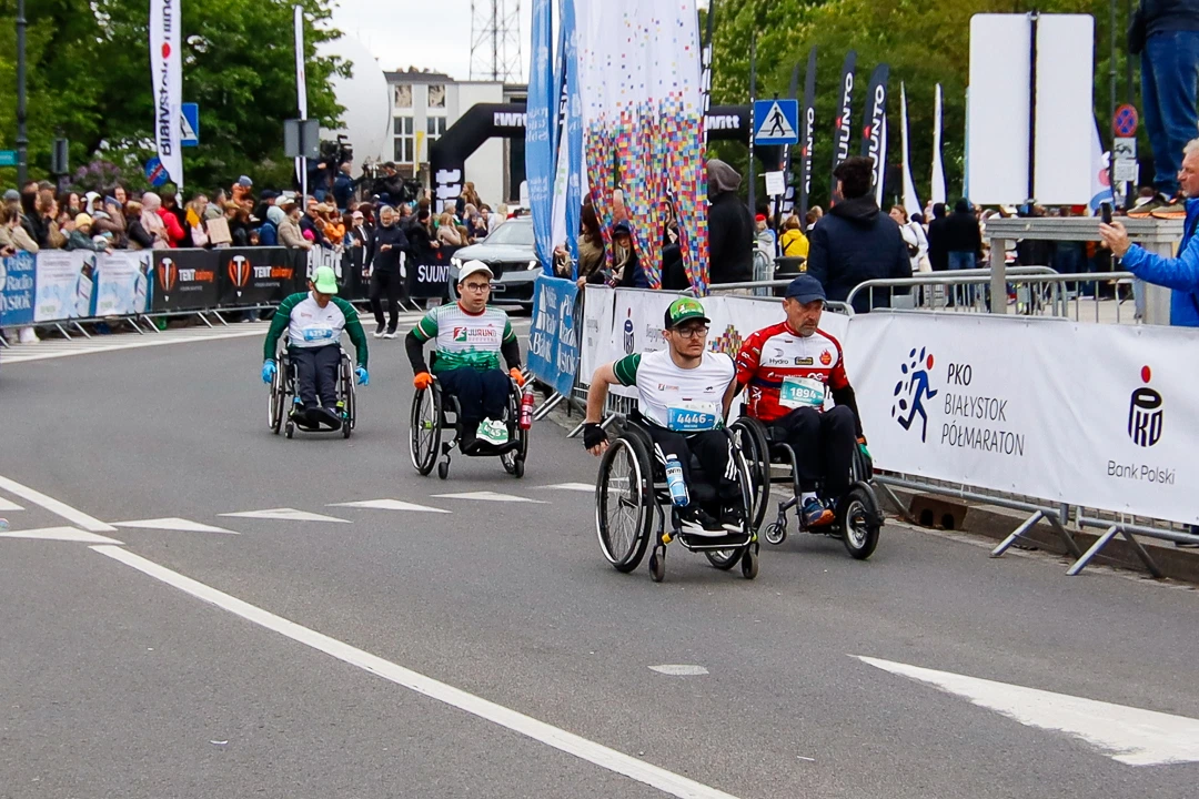 Uczestnicy na wózkach przy banerach PKO Białystok Półmaratonu i kibicującej publiczności