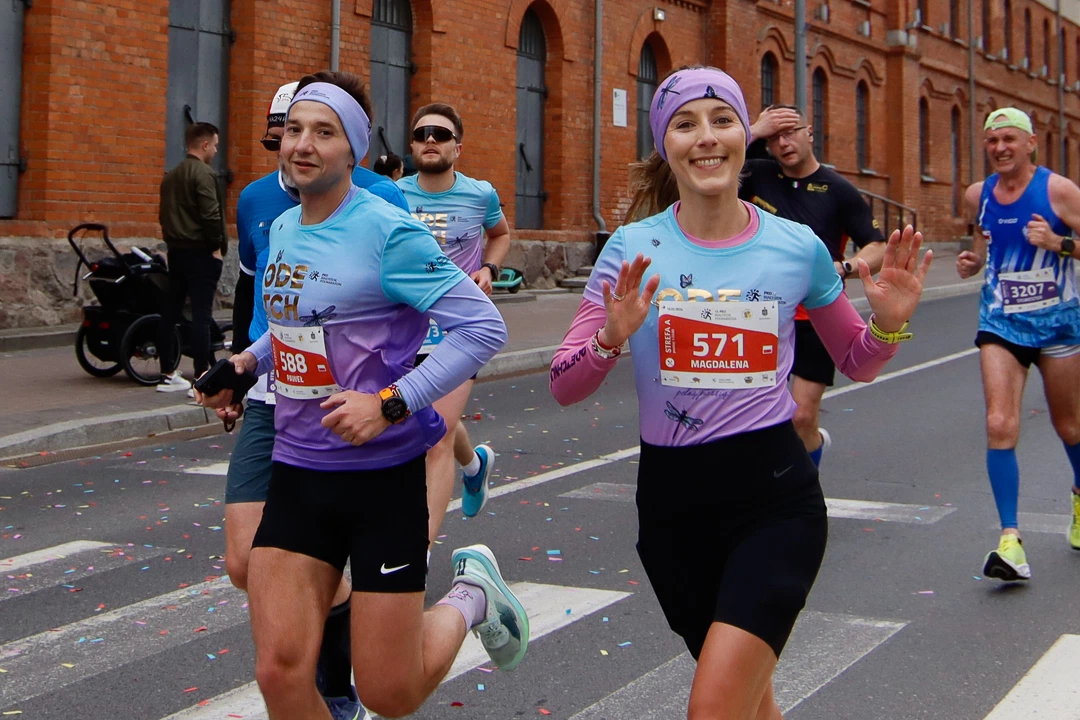 Uśmiechnięci uczestnicy w turkusowo-fioletowych koszulkach na trasie półmaratonu