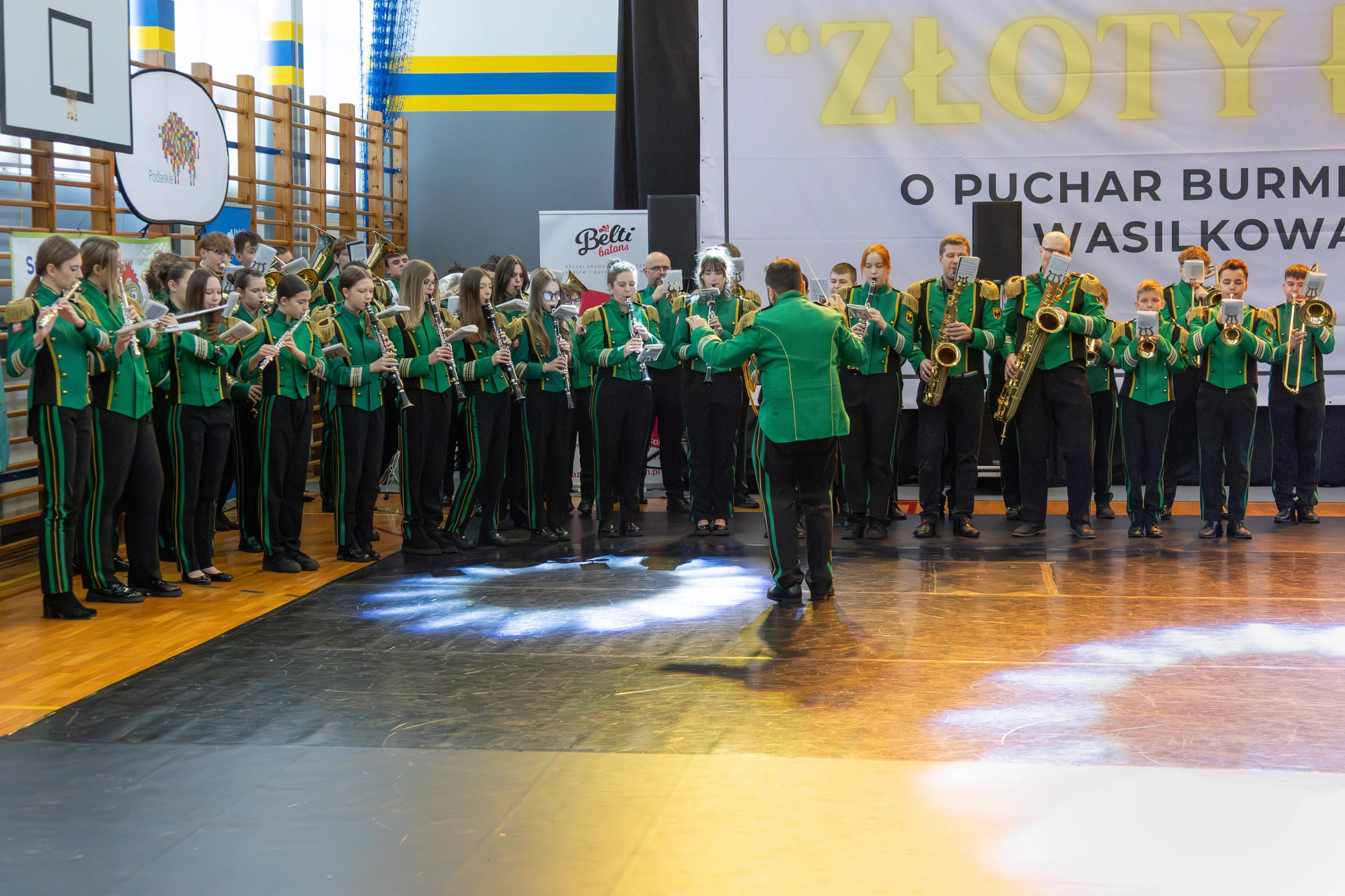 Orkiestra w zielonych mundurach grająca pod kierunkiem dyrygenta przy banerze turnieju