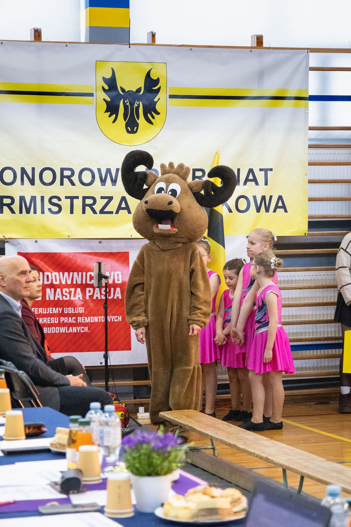 Maskotka łosia stojąca obok uczestniczek przy banerze z herbem Wasilkowa